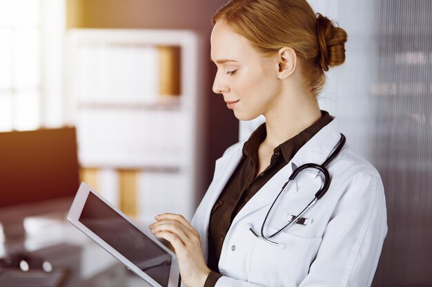 Medico femminile sorridente allegro che utilizza il computer della compressa nella clinica soleggiata. Ritratto di donna medico amichevole al lavoro. Concetto di medicina.