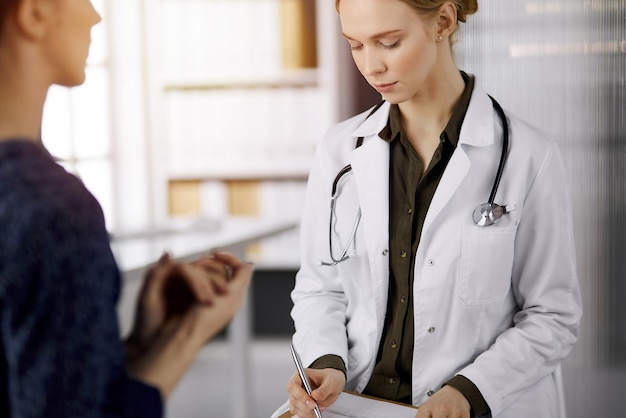 Medico femminile sorridente allegro e donna paziente che discutono l'esame di salute corrente mentre si siedono nella clinica soleggiata. Servizio medico perfetto in ospedale. Concetto di medicina.