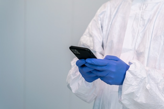 Medico in DPI e utilizzo di guanti per le mani che consulta l'applicazione mobile su smartphone durante una pandemia di covid 19. Operatore sanitario in dispositivi di protezione individuale.