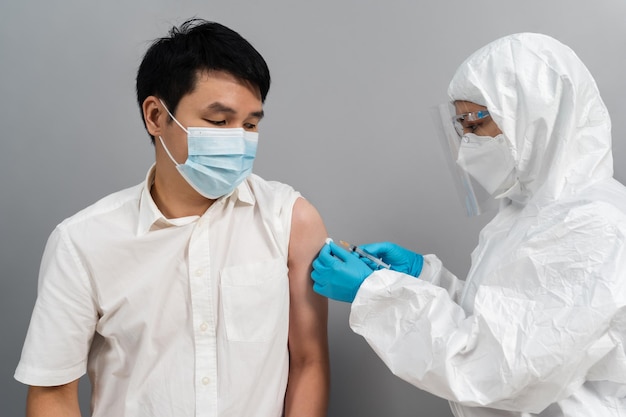 Medico in siringa per tuta protettiva DPI e utilizzo di cotone prima di effettuare l'iniezione al paziente in maschera medica. Vaccino Covid-19 o coronavirus
