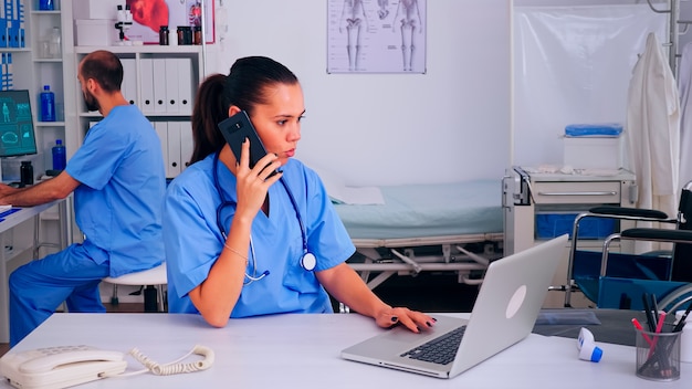 Medico infermiere che consulta paziente remoto utilizzando lo smartphone in ospedale indossando l'uniforme medica. Assistente medico medico che aiuta il paziente con la comunicazione di telemedicina, la diagnosi, la digitazione sul laptop