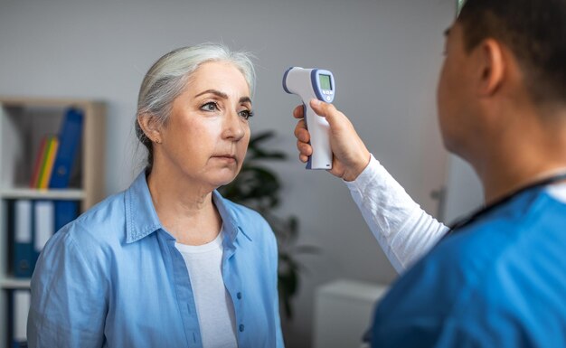 Medico maschio asiatico adulto che controlla la temperatura con il termometro senza contatto al paziente anziano triste della signora