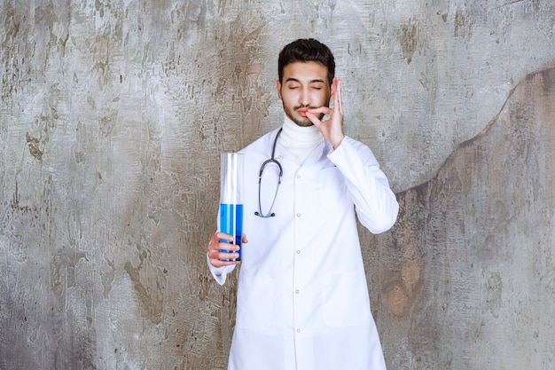 Medico maschio con lo stetoscopio che tiene una boccetta chimica con liquido blu all'interno e che mostra il segno riuscito della mano.
