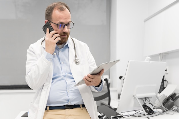 Medico maschio con stetoscopio seduto alla scrivania che parla sul telefono cellulare e guarda la tavoletta digitale che discute di una diagnostica del paziente