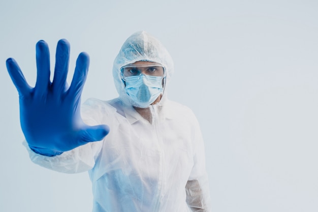 Medico maschio europeo che mostra gesto di arresto. Uomo che indossa camice bianco, occhiali, maschera protettiva e guanti in lattice. Concetto di lotta al coronavirus. Sfondo grigio con luce turchese. Copia spazio