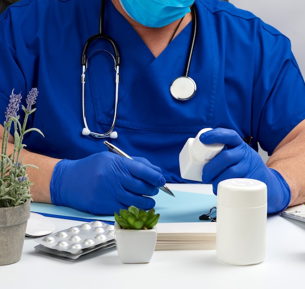 Medico maschio in uniforme blu si siede a un tavolo bianco e scrive su un quaderno di carta, indossando guanti sterili sulle mani