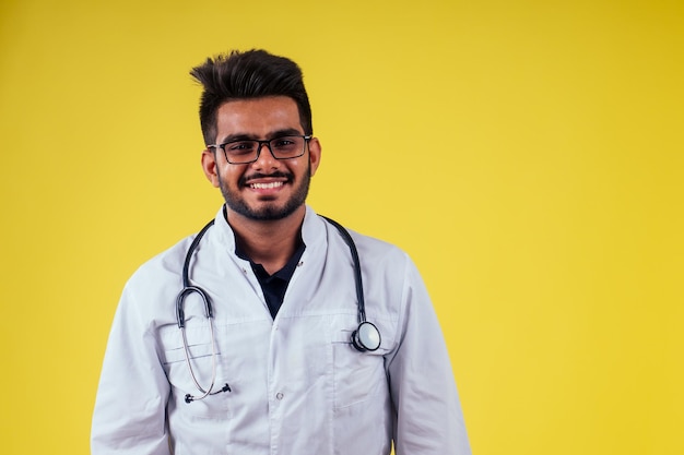 Medico maschio indiano che indossa occhiali e mantello bianco in studio di sfondo giallo.