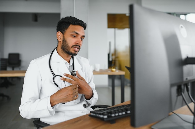 Medico maschio indiano con gli occhiali camice medico bianco e stetoscopio seduto alla scrivania con il computer portatile in una clinica moderna e videoconferenze online coinvolte consulenze da remoto