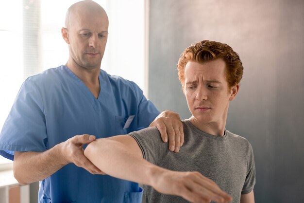 Medico maschio maturo in uniforme blu che sostiene il braccio piegato di un giovane paziente durante l'allenamento fisioterapico nelle moderne cliniche di riabilitazione