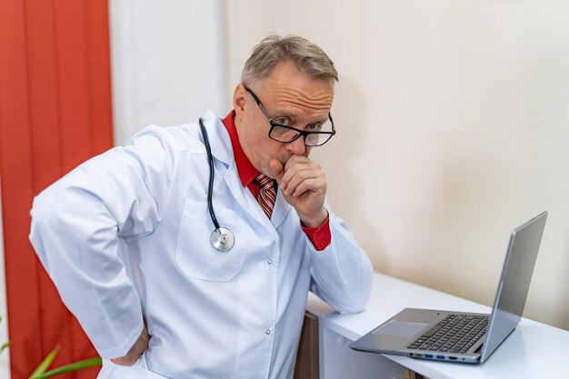 Medico maturo premuroso con il computer portatile sulla scrivania in clinica Medic pensa alla cura futura di un paziente