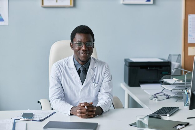 Medico nero sorridente che guarda l'obbiettivo seduto alla scrivania nell'ufficio della clinica