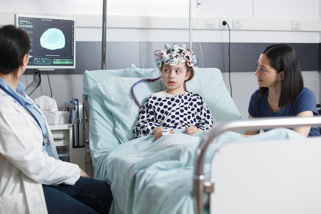 Medico pediatrico che analizza i risultati della scansione dell'elettroencefalogramma di una bambina malata mentre si trova nella sala di risveglio del reparto pediatrico. Paziente bambino che indossa un casco per elettroencefalografia mentre è in clinica pediatrica