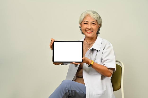 Medico senior amichevole della donna in camice bianco che tiene compressa digitale che si siede contro il fondo marrone