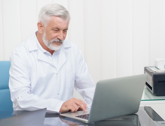 Medico senior che indossa in uniforme bianca che lavora con il computer portatile.