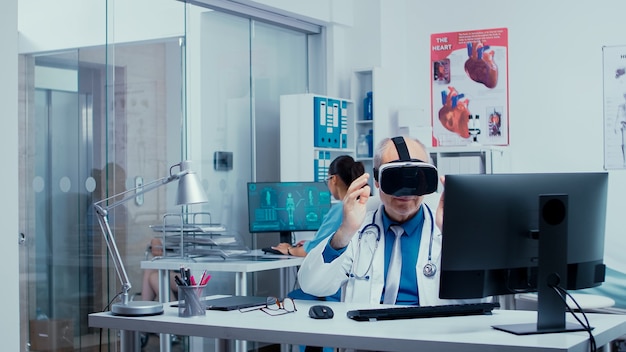 Medico senior che utilizza occhiali VR in una moderna clinica privata per studiare le malattie nello spazio virtuale e la tecnologia moderna. Sullo sfondo una clinica moderna con pareti di vetro e pazienti con medici in hallw