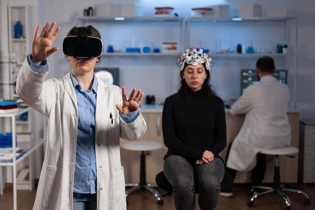 Medico specialista neurologo con auricolare per realtà virtuale che analizza l'evoluzione del cervello durante l'esperimento di neuroscienze in laboratorio medico. Paziente donna con scanner EEG affetta da malattia neurologica