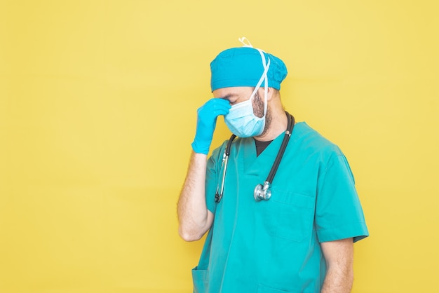 Medico vestito da chirurgo in verde con stetoscopio e maschera su sfondo giallo con espressione preoccupata.