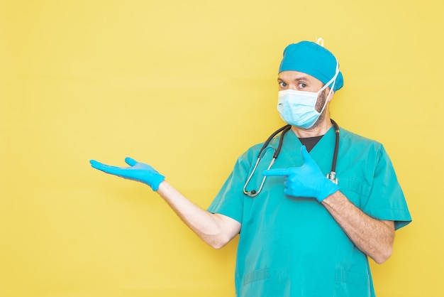 Medico vestito da chirurgo in verde con stetoscopio e maschera su sfondo giallo con gesto che punta a una direzione.