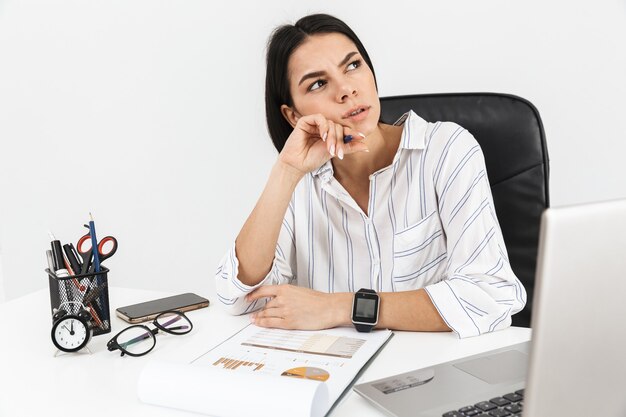 meditabondo bruna imprenditrice lavorando con documenti cartacei mentre era seduto al tavolo in ufficio isolato sopra il muro bianco