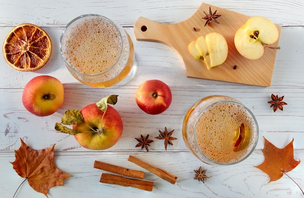 Mele rosse succose fresche mature, spezie e bicchieri con sidro su una superficie in legno chiaro.