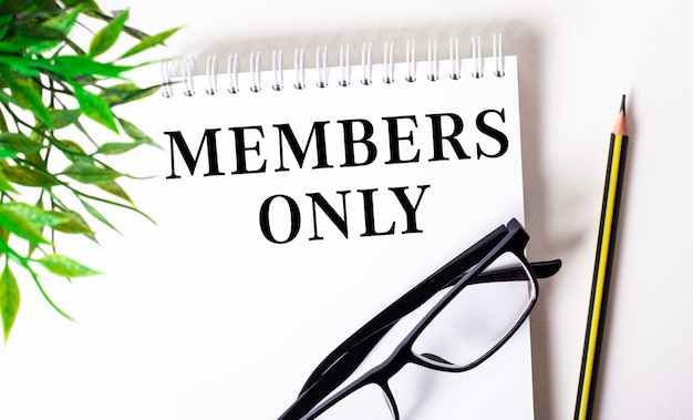 MEMBERS ONLY è scritto su un quaderno bianco accanto a una matita, occhiali con cornice nera e una pianta verde