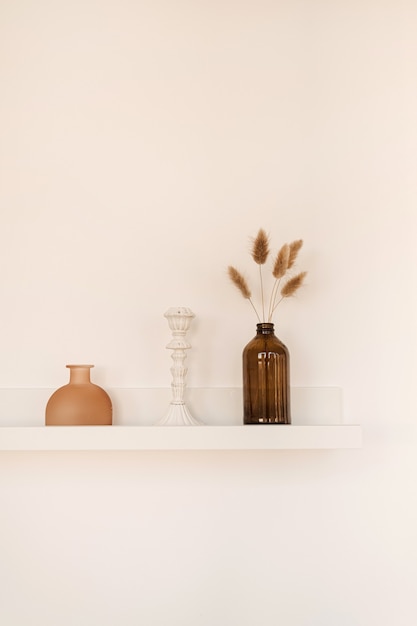 Mensola bianca sul muro bianco con bottiglia, vaso, soffici piante di pom pom, candeliere.