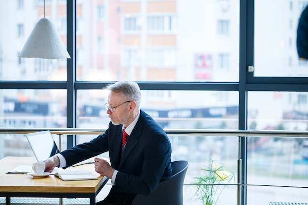 Mentore maschio adulto, direttore, uomo d'affari con gli occhiali e un vestito che studia documenti mentre è seduto al tavolo. Concetto di giornata lavorativa