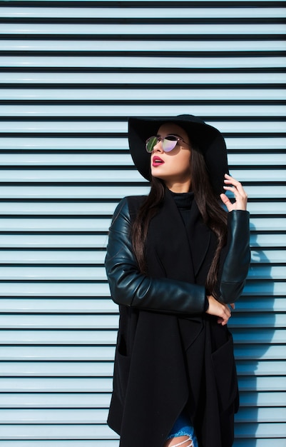 Meravigliosa donna bruna in elegante cappotto e cappello in posa vicino all'otturatore in una giornata di sole.