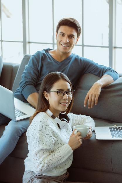 Mescola gli amanti della famiglia razziale, il marito caucasico e la moglie asiatica, seduti in soggiorno a lavorare tramite computer portatile sul divano in semplici gesti di relax. Concetto di lavoro a casa e nuovo stile di vita moderno normale.