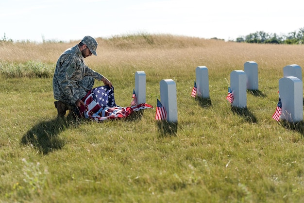 Messa a fuoco selettiva del soldato in uniforme e berretto con bandiera americana mentre si siede nel cimitero