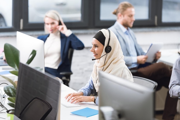 messa a fuoco selettiva dell'operatore sorridente del call center arabo in hijab in cuffia che lavora sul computer