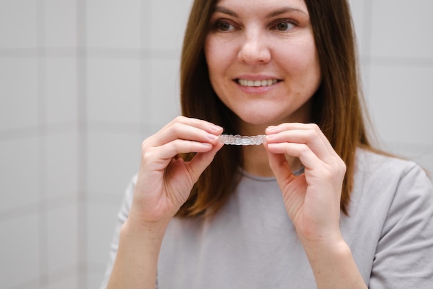 Messa a fuoco selettiva Primo piano di una donna sorridente davanti a uno specchio in bagno che tiene in mano allineatori di denti in plastica invisibili pronti a mettere le parentesi graffe Sorriso bello e sano