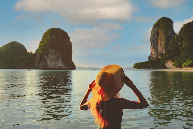 Messa a fuoco selettiva sul cappello di paglia giallo sul retro della donna in piedi sulla spiaggia durante il tramonto al tropicale Railay Beach Krabi Thailandia