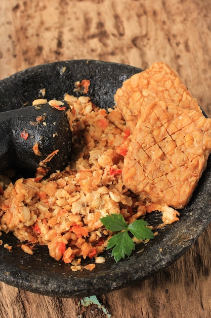 Messa a fuoco selezionata Sambal Tempeh, Tempe schiacciato piccante su pestello di pietra. Cibo tradizionale indonesiano con gusto piccante e salato