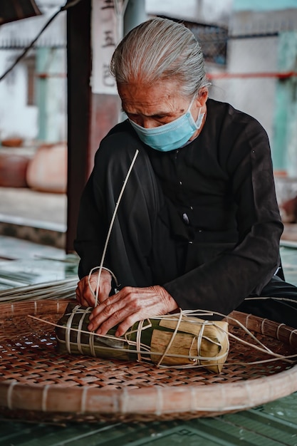 Messa a fuoco vecchia donna con il tradizionale abito vietnamita ao ba ba confezionamento torta Tet il nuovo anno lunare vietnamita Tet cibo all'aperto a mano