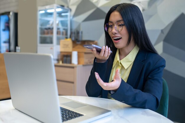 Messaggio audio. Giovane donna asiatica con gli occhiali che parla nel microfono dello smartphone gesticolando seduto al computer portatile in ufficio