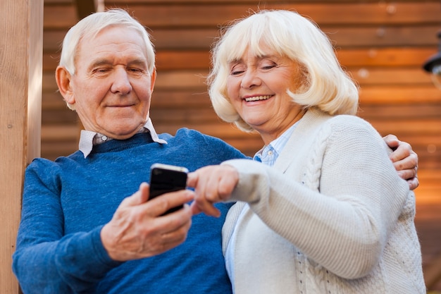 Messaggio dal figlio. Felice coppia di anziani che si legano l'un l'altro e sorridono mentre si trovano all'aperto e guardano il telefono cellulare