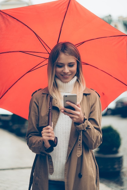 Messaggio veloce in movimento. Attraente giovane donna sorridente che porta ombrello e guarda il suo telefono cellulare mentre cammina per strada