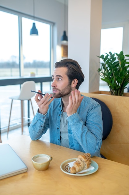 Messaggio vocale. Giovane uomo adulto che guarda lontano parlando tenendo lo smartphone all'altezza della bocca seduto al tavolo nella caffetteria durante il giorno