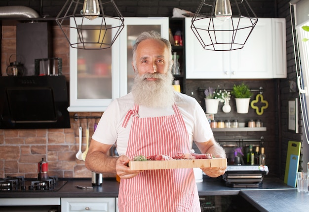 Metà colpo di chef maturo di smiley con tagliere in mano. Carne sdraiata sul tavolo. Uomo isolato in cucina moderna ben organizzata.