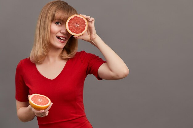 Metà del pompelmo Donna bionda dai capelli raggiante positiva che tiene metà del delizioso pompelmo all'occhio