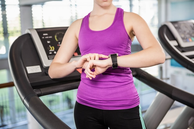 Metà di sezione della donna adatta che utilizza smartwatch sul tapis roulant in palestra