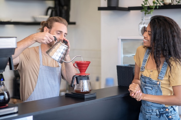 Metodo di preparazione. Felice barista maschio in piedi dietro il bar, fare il caffè con riverso e guardare la donna dai capelli lunghi