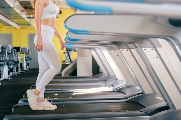 Metta in mostra la donna che corre sul tapis roulant in palestra, si mantiene in forma, brucia calorie sulla macchina da corsa, indossa abbigliamento sportivo bianco e scarpe da ginnastica.