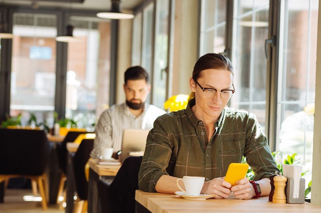 Mezzi di comunicazione. Positivo felice uomo seduto al tavolo del bar mentre manda un sms sul suo smartphone