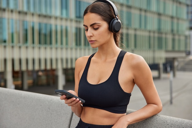 Mezzo busto di modello femminile sportivo motivato scarica app per lo sport ascolta i tipi di musica preferiti sul telefono vestito con canotta utilizza pose di applicazioni yoga nel centro della città