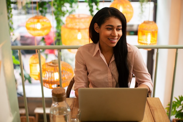 Mezzo busto di un giovane studente soddisfatto seduto al bar da solo e guardando in lontananza con un sorriso mentre ha un laptop moderno di fronte a lei