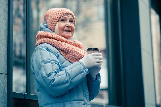 Mezzo busto di una premurosa signora anziana in abiti invernali in piedi con una tazza di caffè in mano e guardando lontano