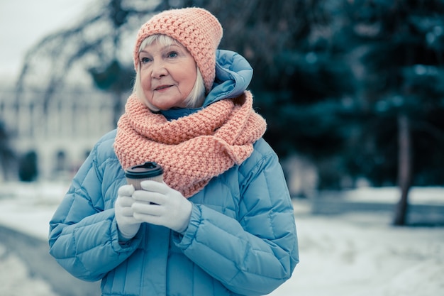 Mezzo busto di una tranquilla vecchia signora in vestiti caldi che sta all'aperto in una giornata invernale e guarda lontano mentre tiene in mano una tazza di caffè in cartone