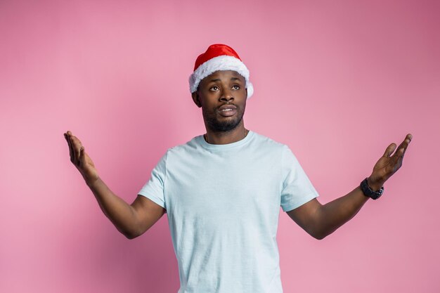 Mezzo busto di uomo afroamericano deluso che allarga le mani con indignazione, indossando il cappello di Babbo Natale, maglietta, alzando lo sguardo perplesso, isolato su sfondo rosa. Emozioni, espressioni, concetto di gesto